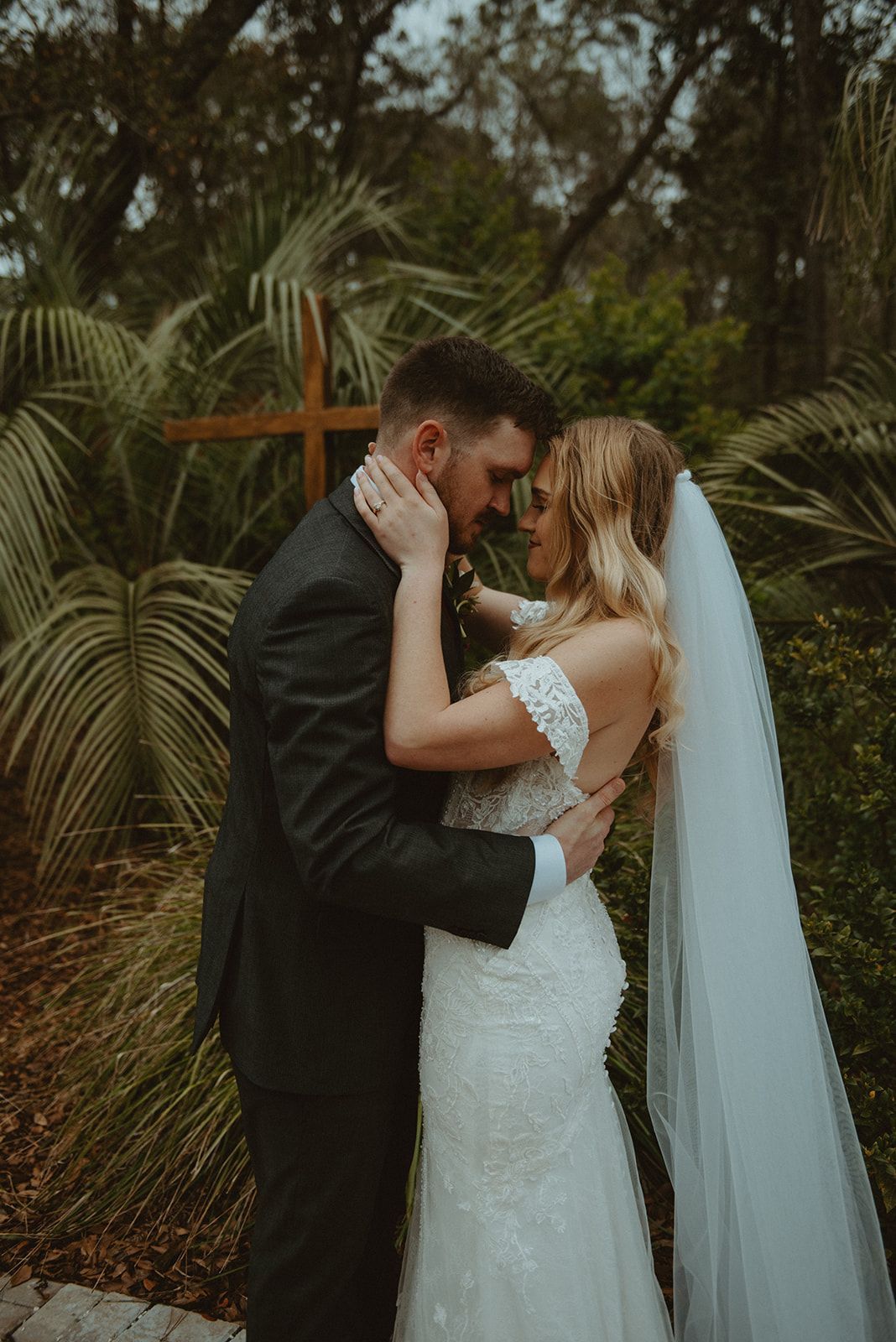 Bride and groom embrace outdoors by a wooden cross, with greenery and a veil flowing behind her.