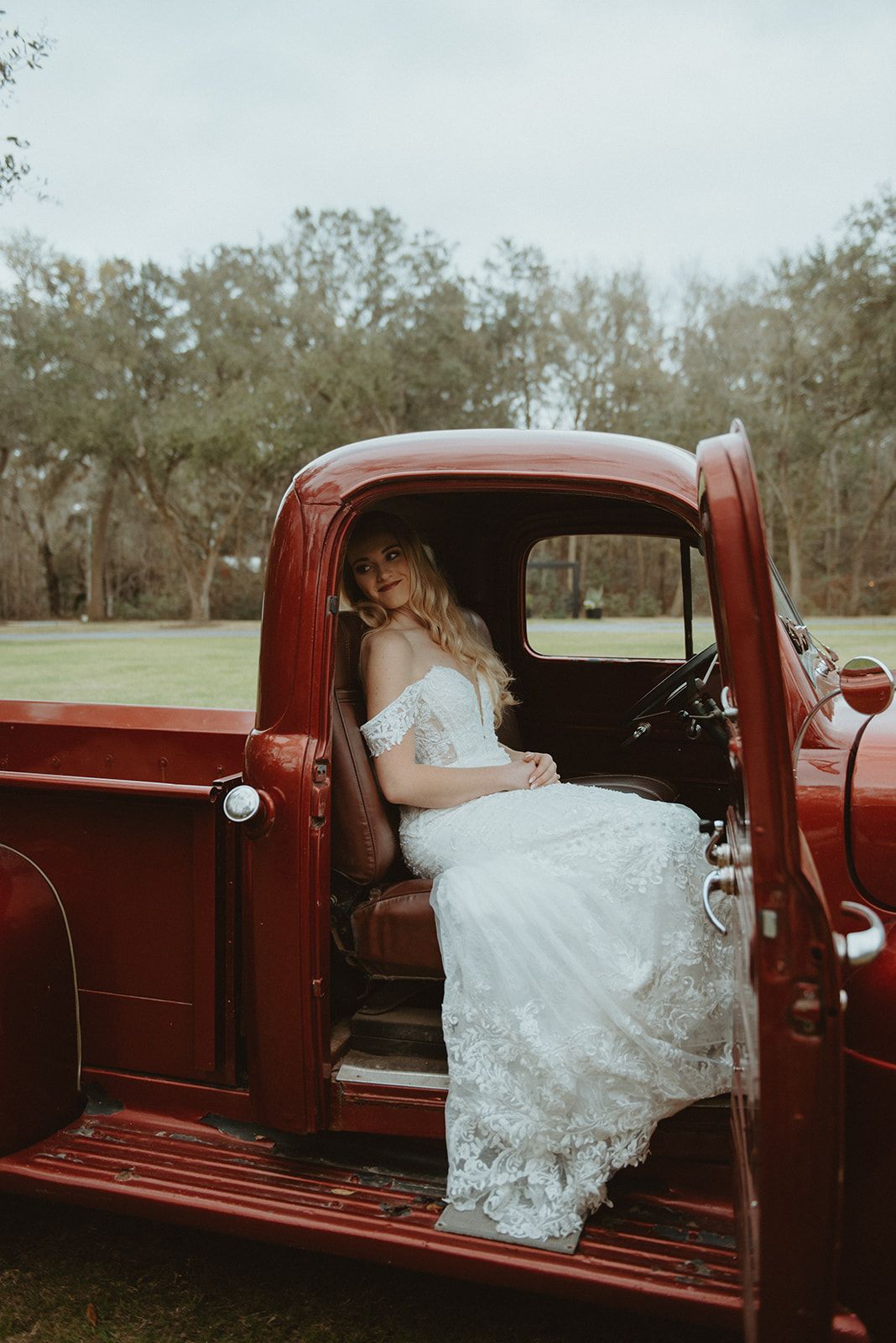 Bride in white lace gown seated in a red vintage truck outdoors with trees in the background