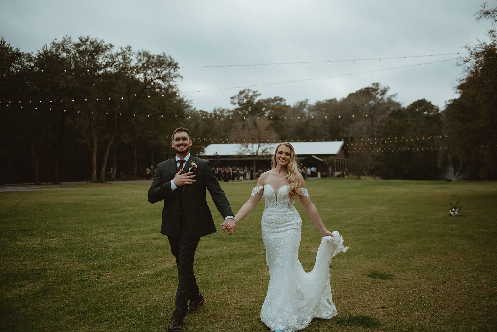 Couple in wedding attire holding hands and walking across a grassy field outdoors.