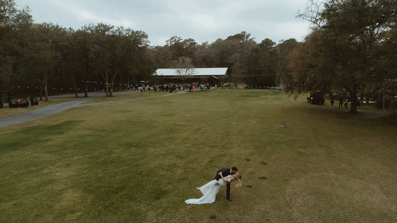 Wide grassy field with a person near a white cloth, trees lining both sides and a pavilion in the distance.