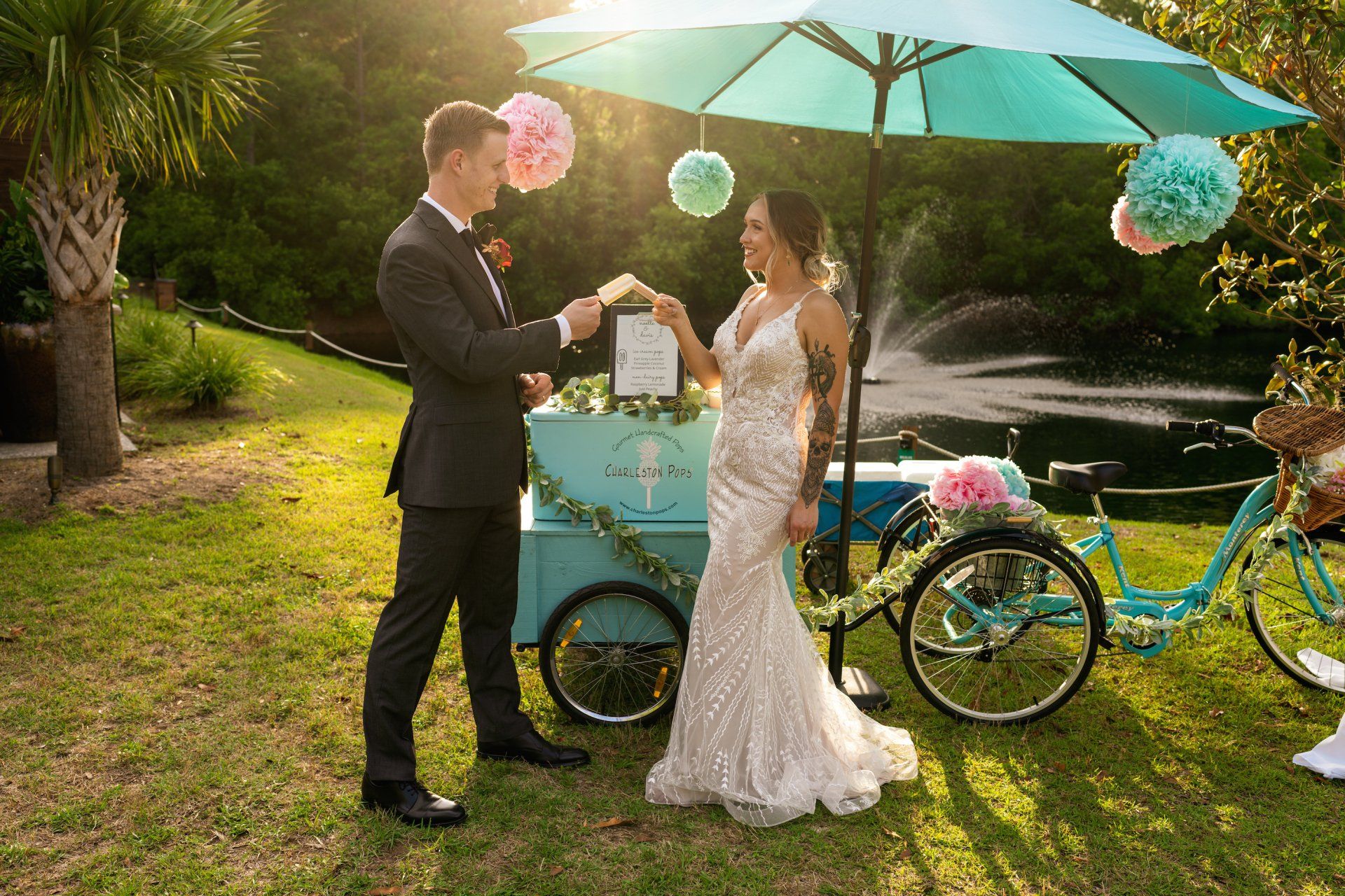 Couple by a turquoise wedding cart under an umbrella, with balloons and a bicycle in a sunny garden