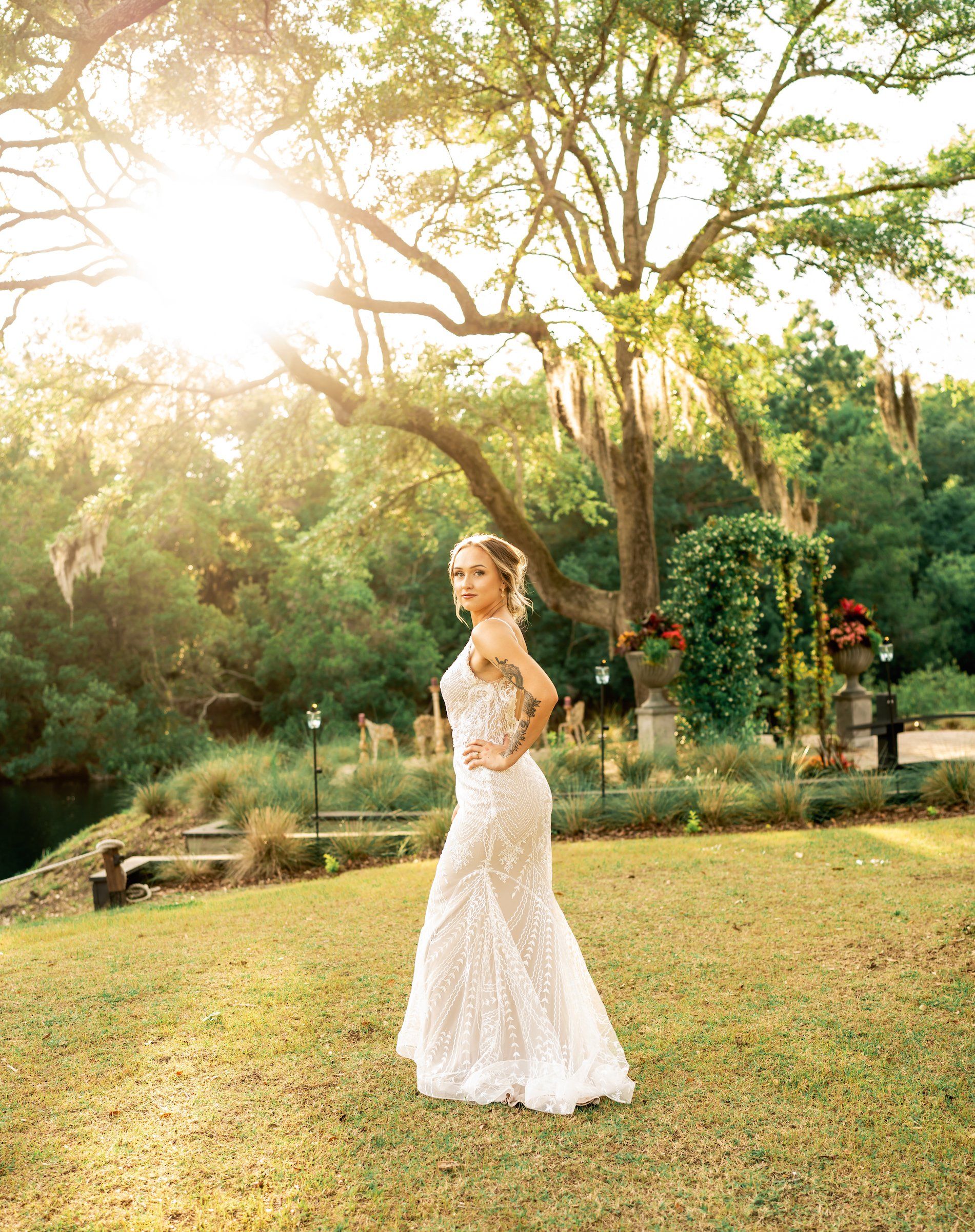 Bride in a white lace gown standing on a sunlit lawn with trees and garden arches behind her