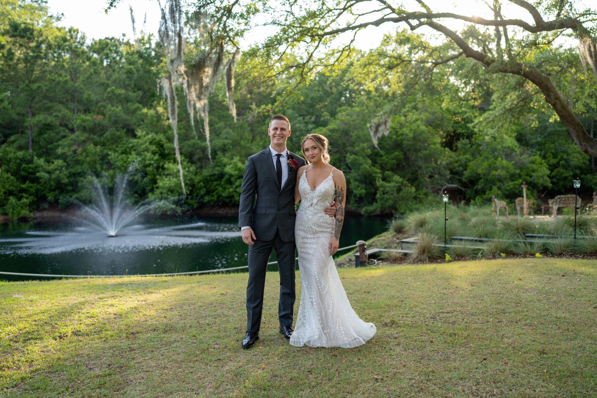 Couple in formal attire standing on a lawn by a pond and fountain in a lush garden at sunset
