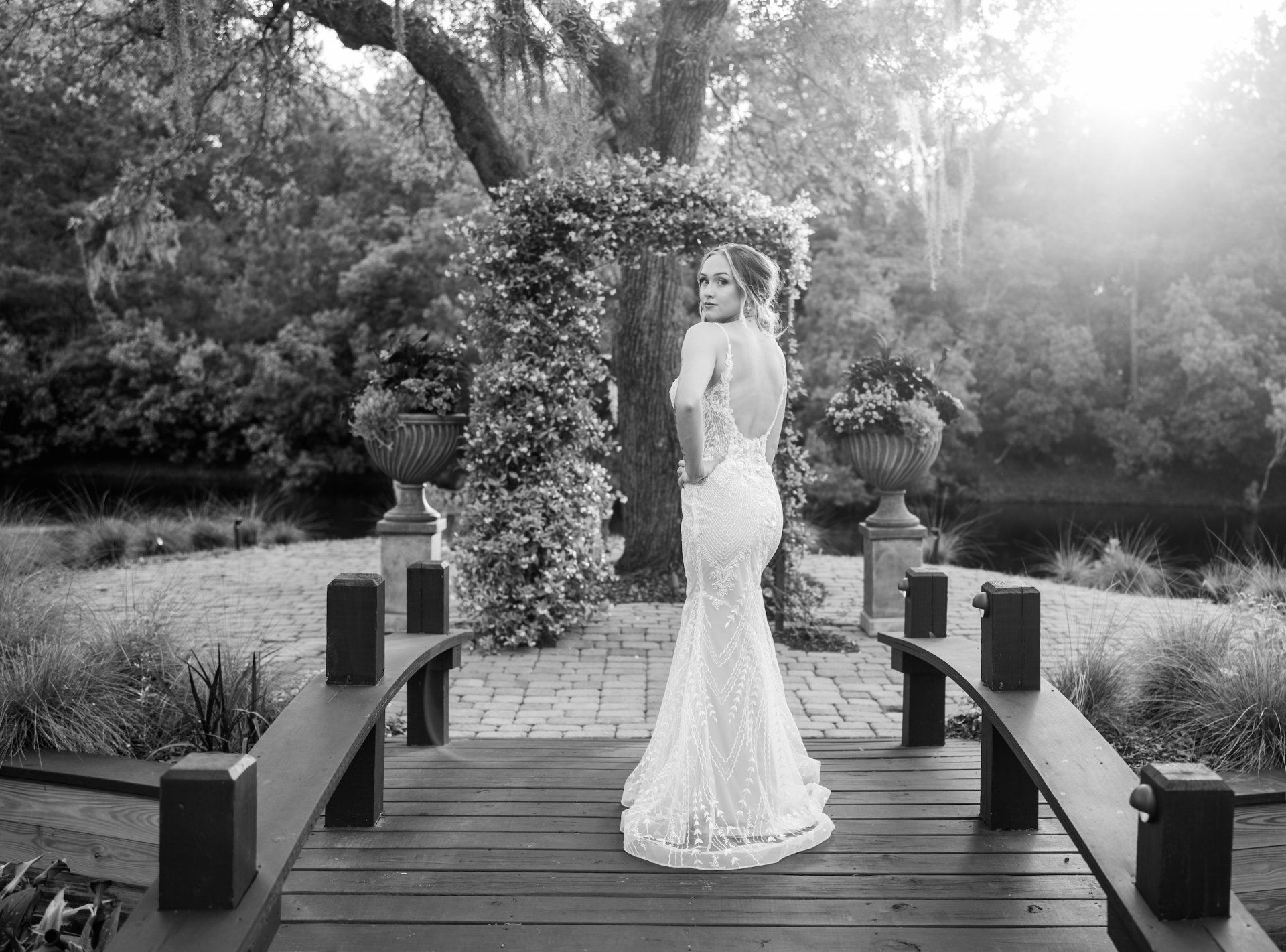 Bride in a white gown standing on a wooden bridge under a floral arch in a garden
