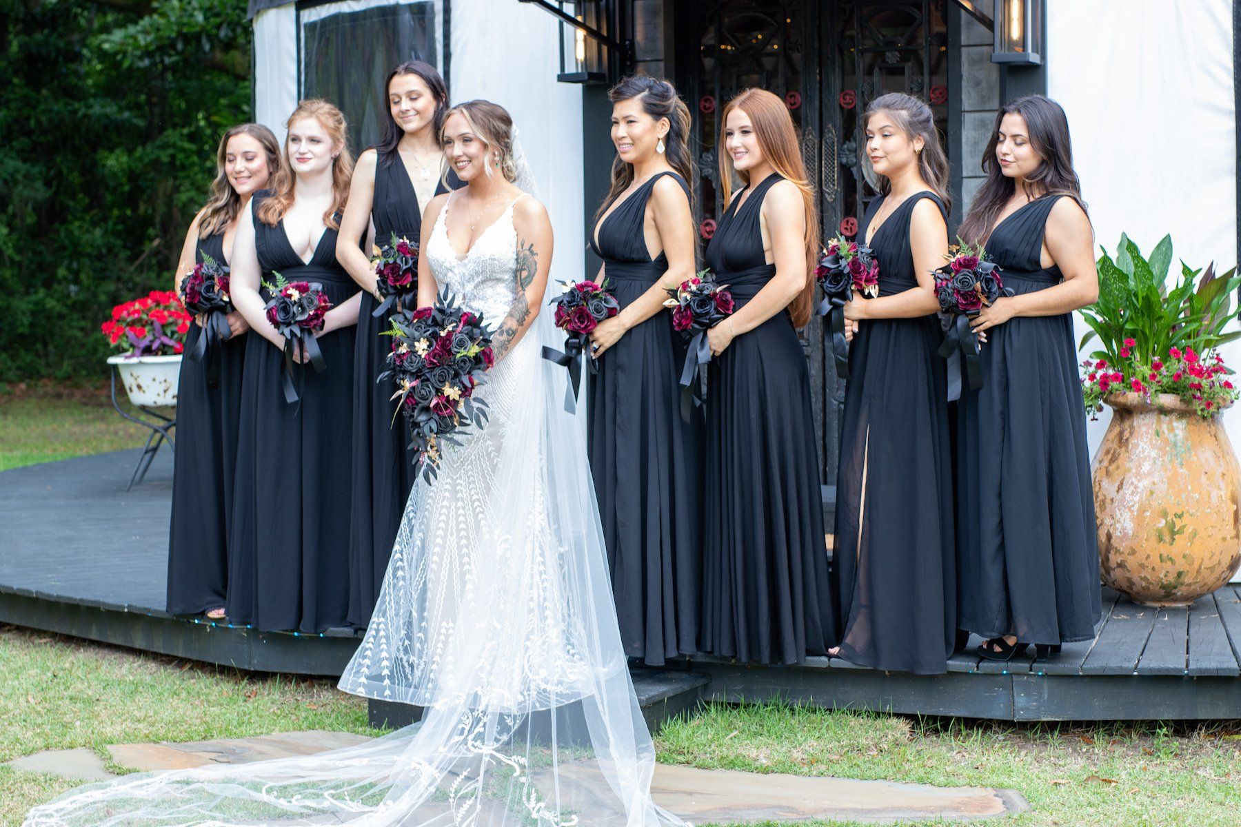 Bride in white gown with black-clad bridesmaids holding bouquets outdoors by a building