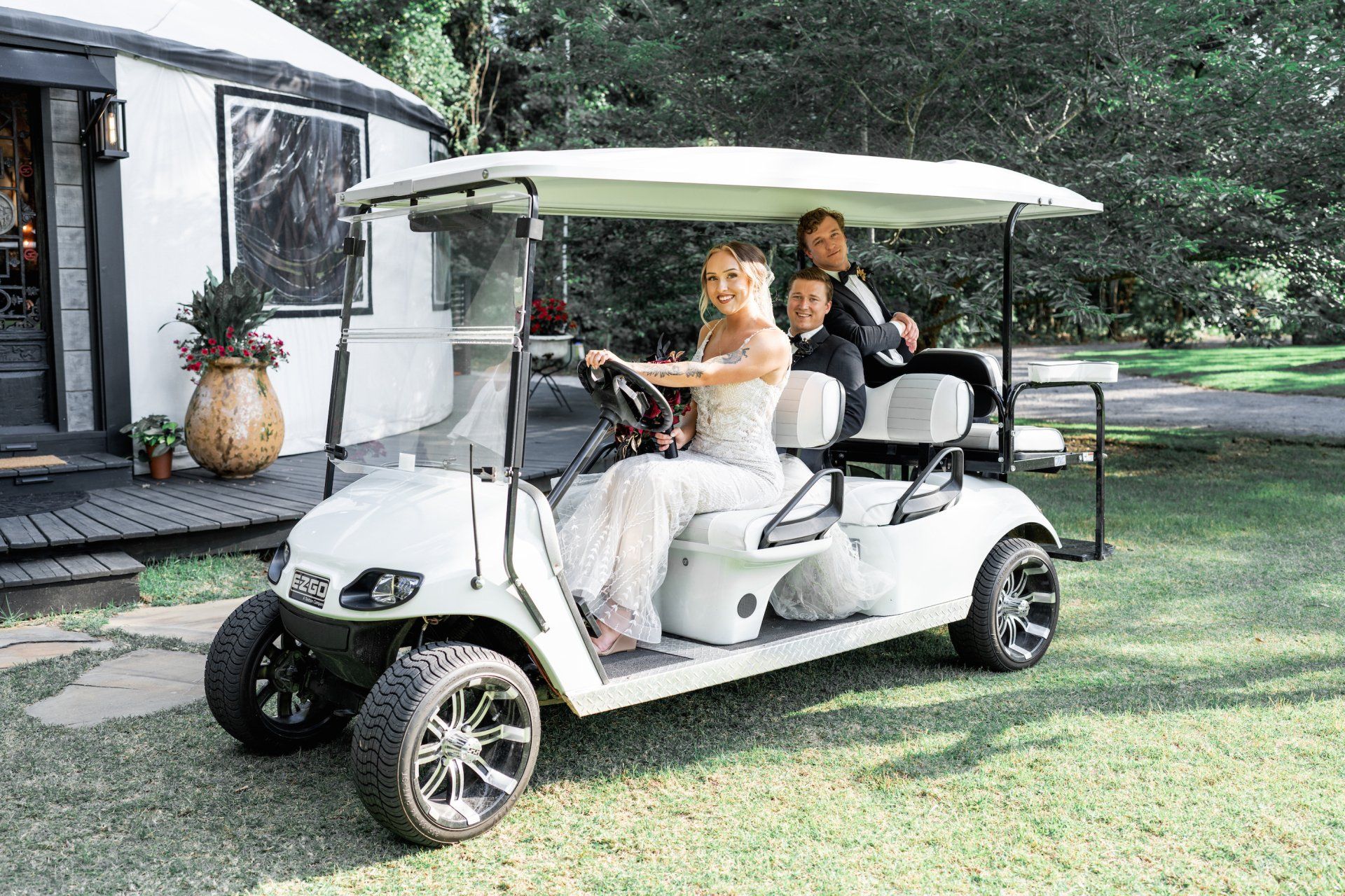 Bride and groom seated in a white golf cart on a lawn near a house, smiling at the camera