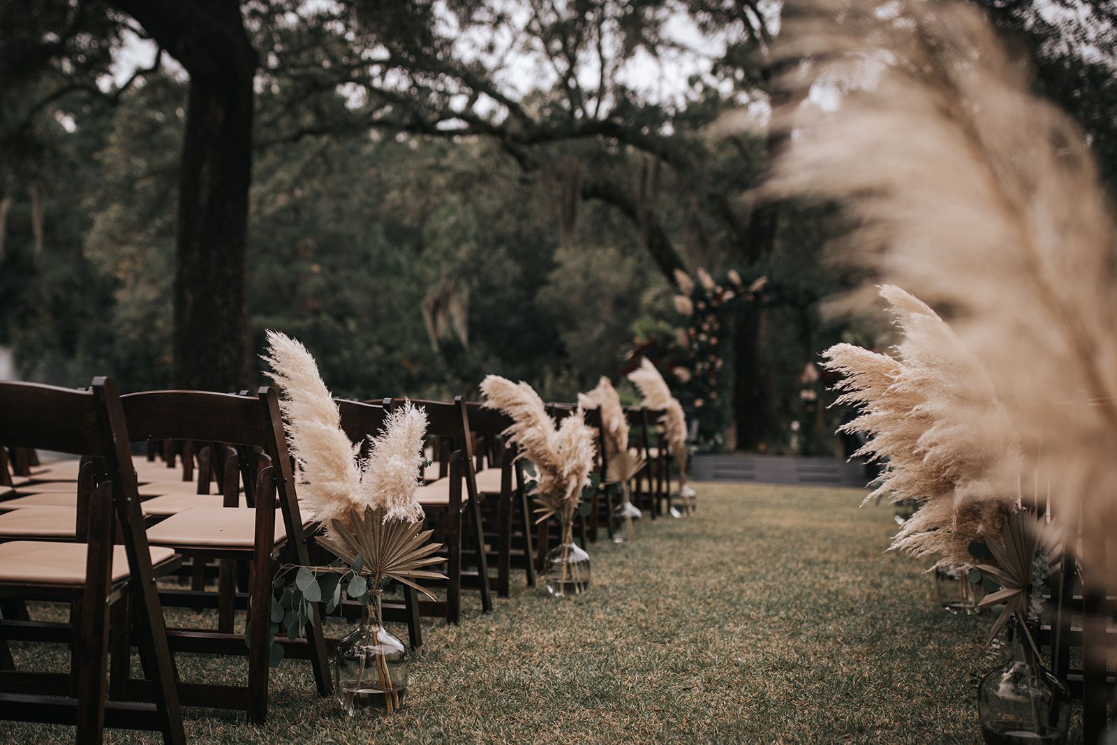 Outdoor wedding setup with wooden chairs and pampas grass along a grassy aisle under trees