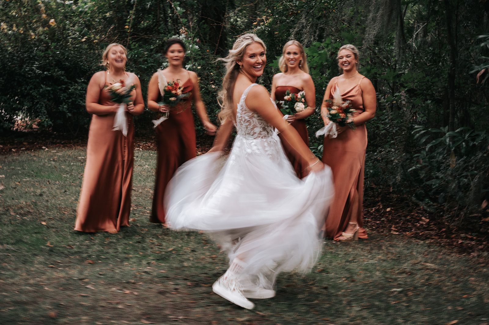 Bride in white dress twirls with four bridesmaids in rust dresses in a wooded outdoor setting