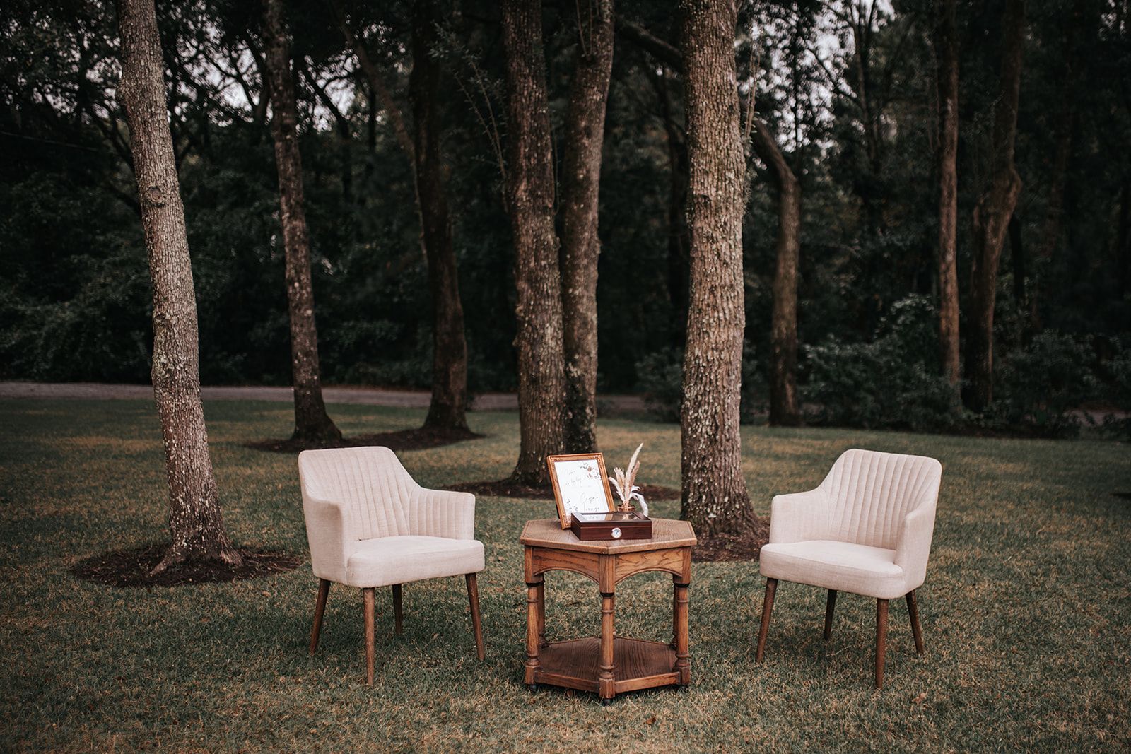 Two white chairs and a small wooden table set on grass in a wooded park clearing.