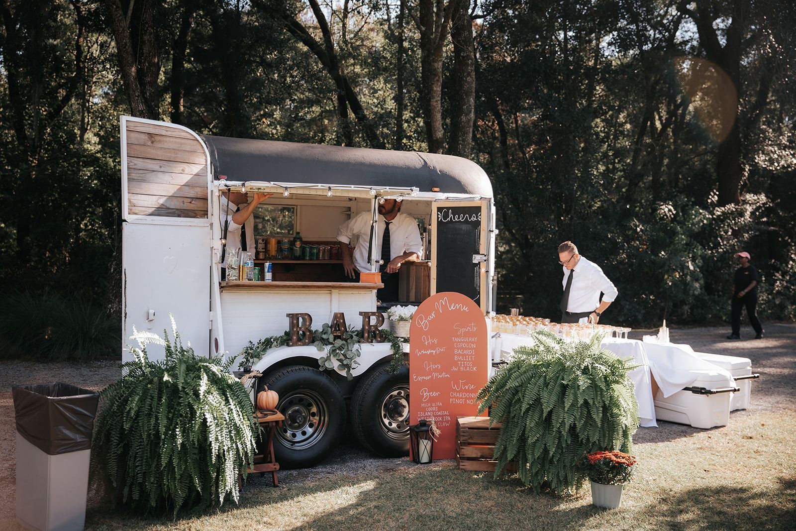 Coffee cart at an outdoor event, with a vendor and white seating under trees