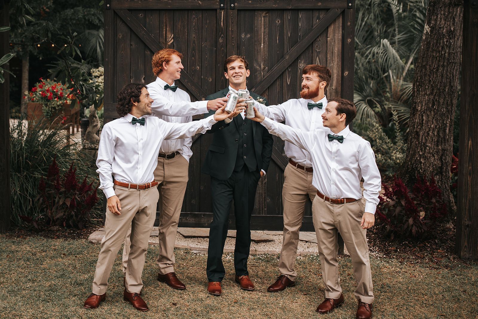 Five people in dress shirts and suspenders toast drinks in front of rustic barn doors, smiling outdoors