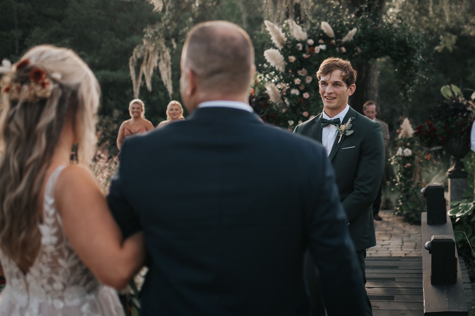 Wedding guests watch as a groom stands smiling outdoors on a wooden walkway.