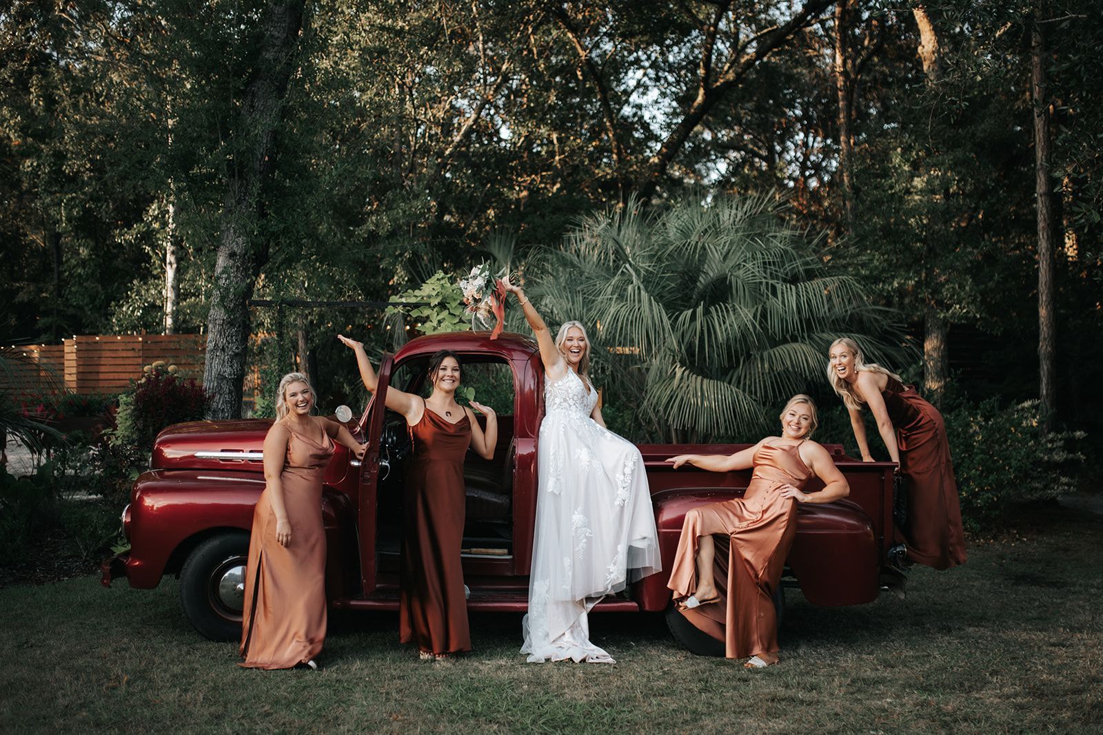 Group posing around a red vintage truck in a wooded outdoor setting, with one person in a white dress waving.