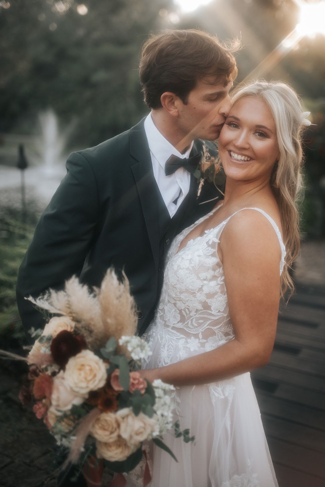 Newlywed couple posing outdoors, bride in white lace dress holding a bouquet, groom kissing her forehead