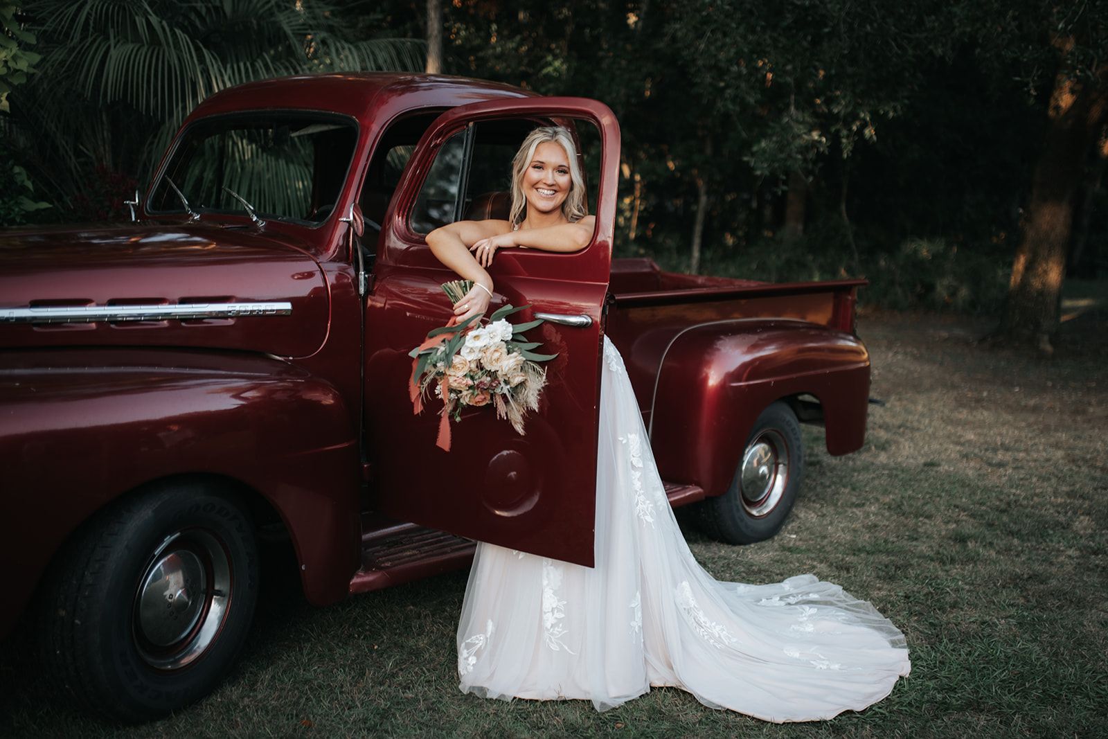 Bride in a white gown leaning from a burgundy vintage pickup truck, holding a bouquet outdoors