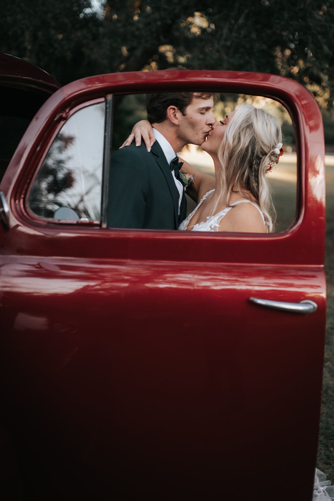 Couple kissing inside a red vintage truck, seen through the open driver-side door.