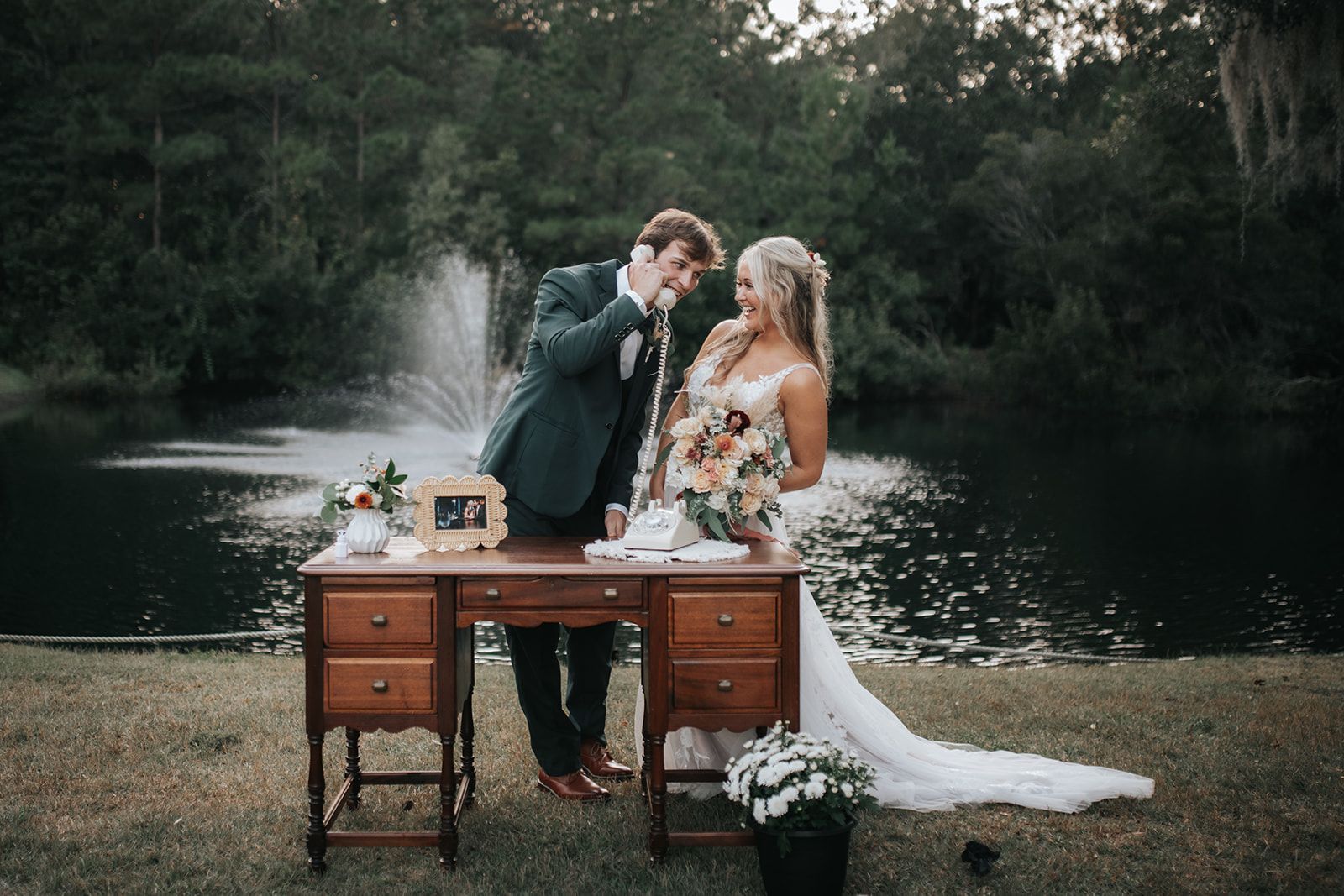 Couple toasting at a rustic outdoor table beside a lake, with a fountain and floral arrangements nearby