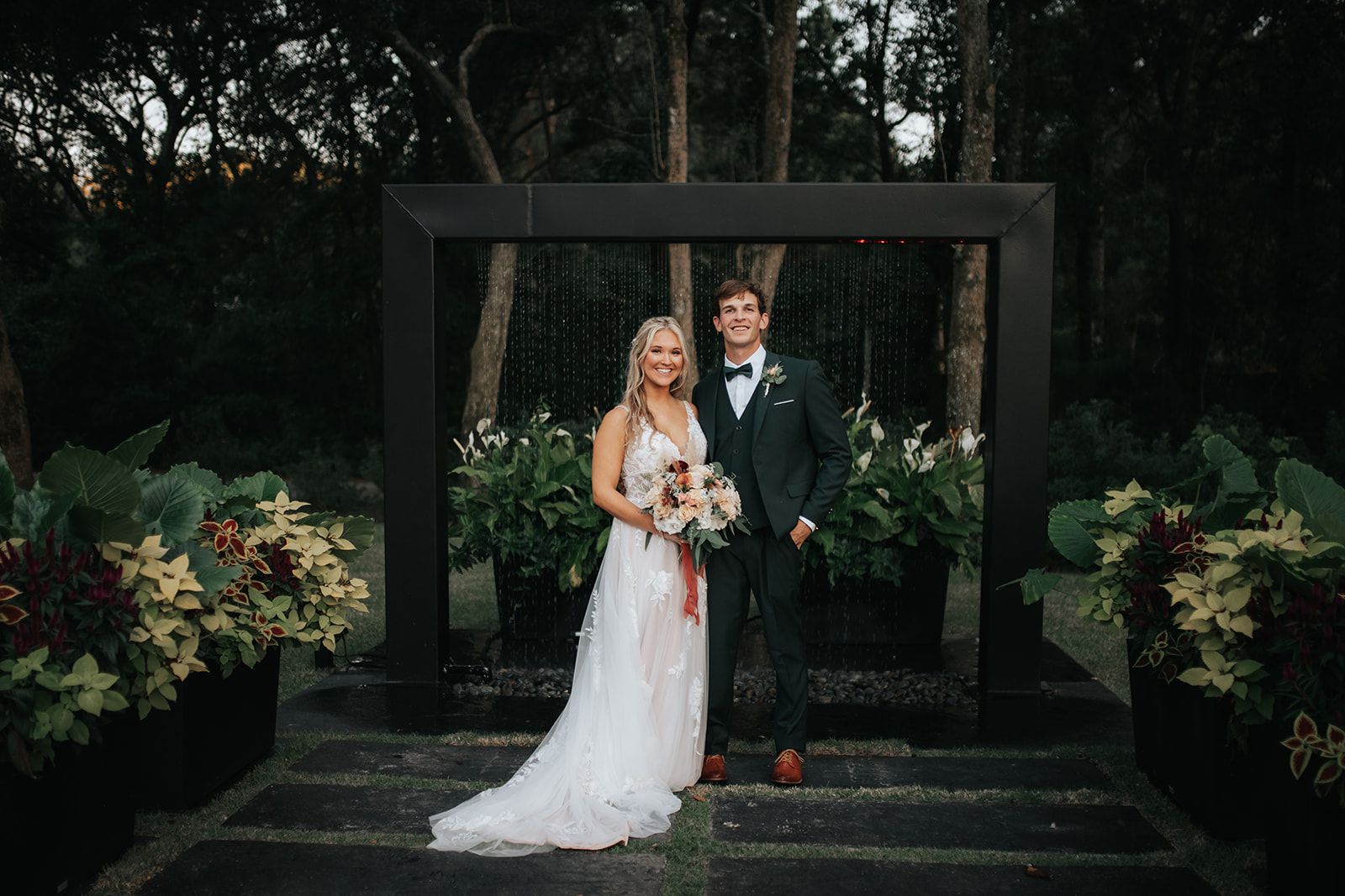 Bride and groom stand together in a dark garden archway, holding a bouquet and smiling.