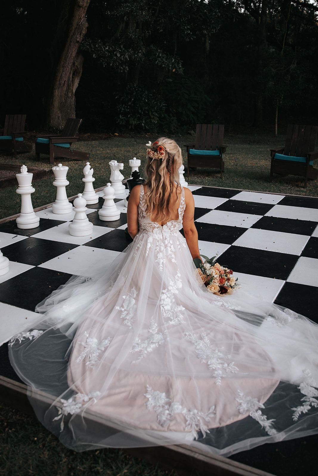 Bride in white gown seated on a giant black-and-white chessboard in an outdoor garden setting