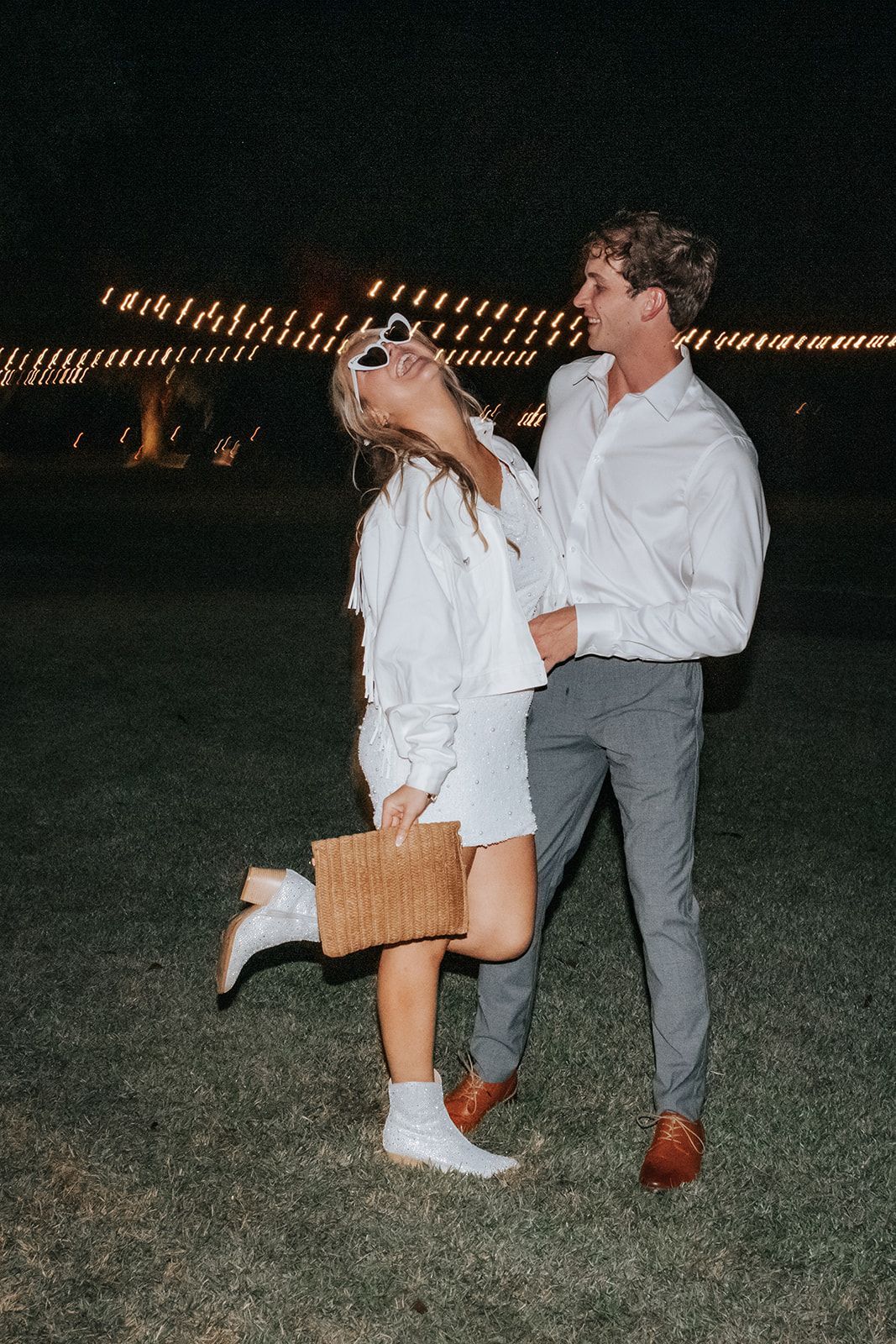 Two people standing on grass at night, one lifting the other while holding hands, with string lights in the background.
