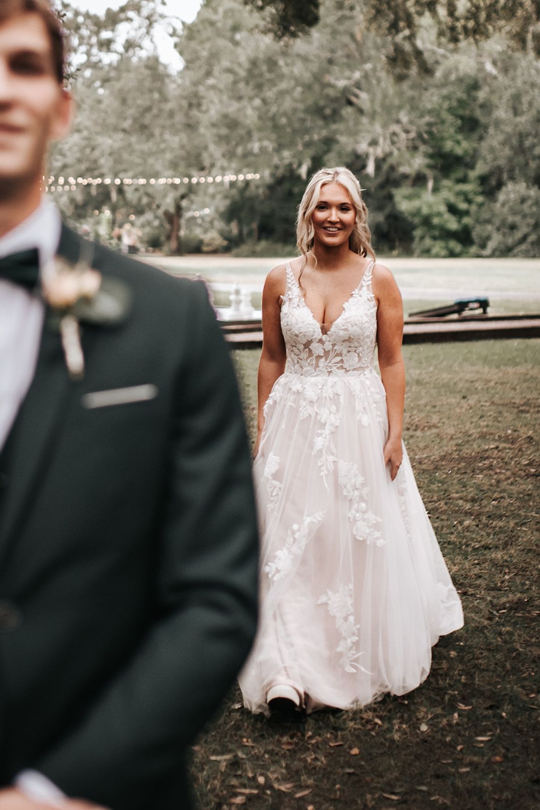 Bride in a white lace gown walking outdoors, with groom blurred in foreground