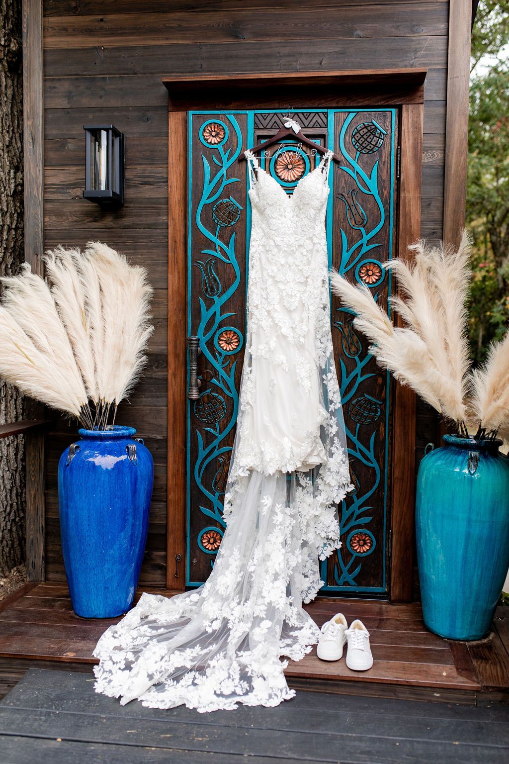 White lace wedding dress hanging on a decorative door, flanked by blue vases with pampas grass.