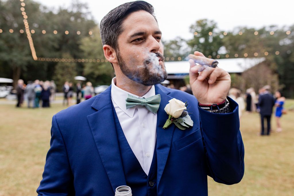 Man in a blue suit smoking an e-cigarette outdoors at a formal event, with guests blurred in the background.