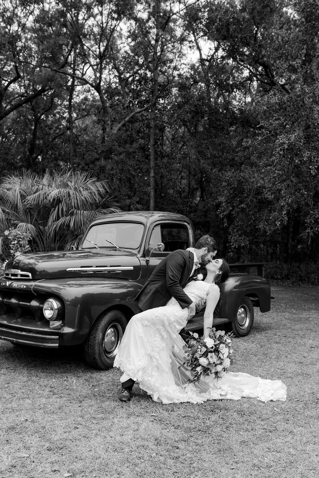 Bride in white gown kneels beside a black vintage truck, bouquet on ground, in a wooded outdoor setting