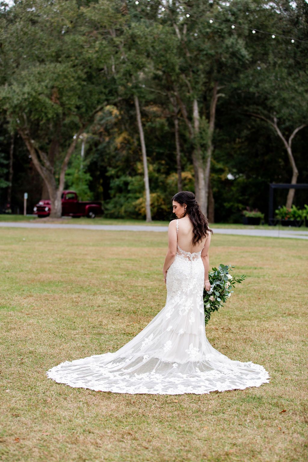Bride in a white lace gown stands in a grassy park, holding greenery and looking over her shoulder.