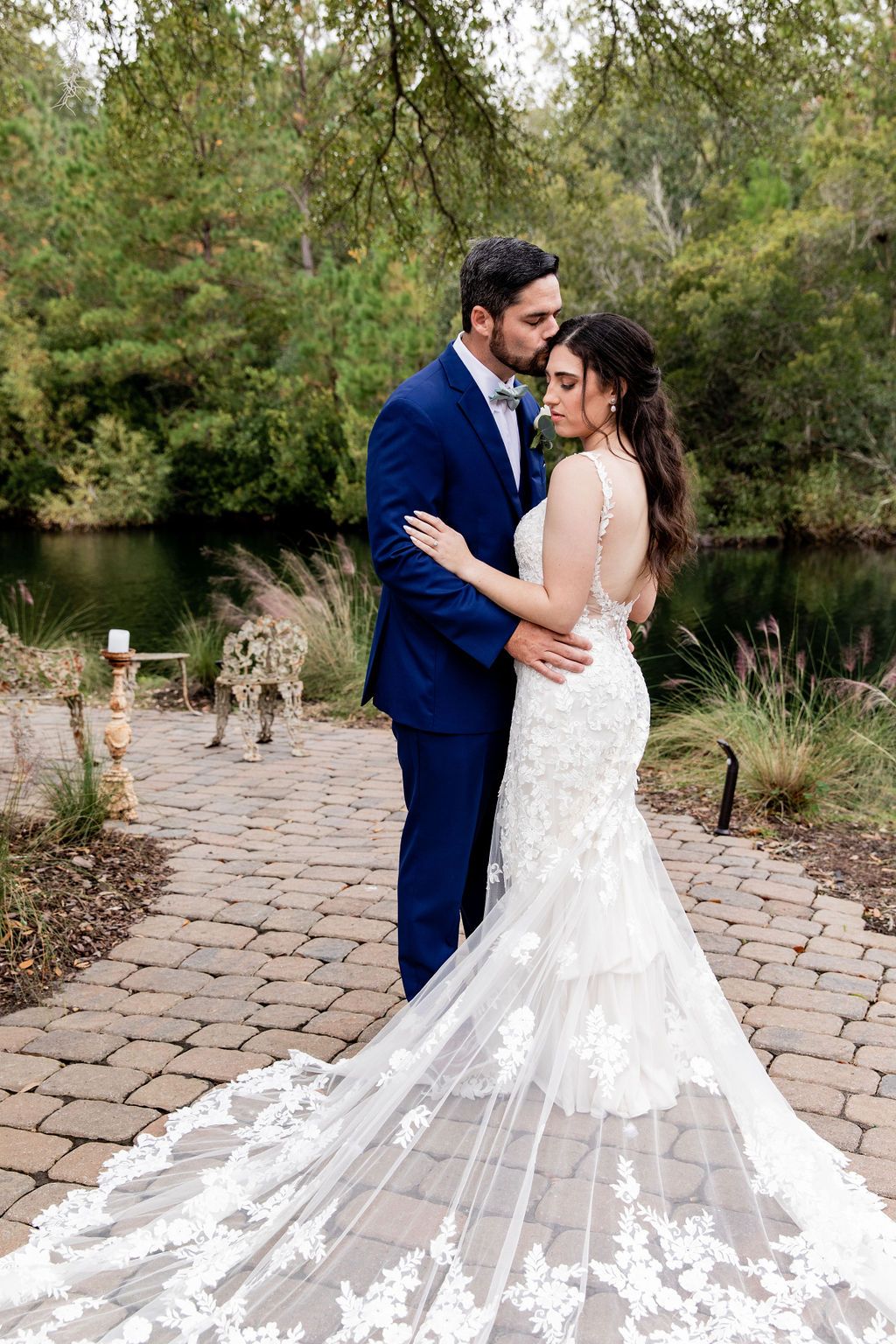 Couple in blue suit and white lace gown embracing on a cobblestone path by a lake and trees.