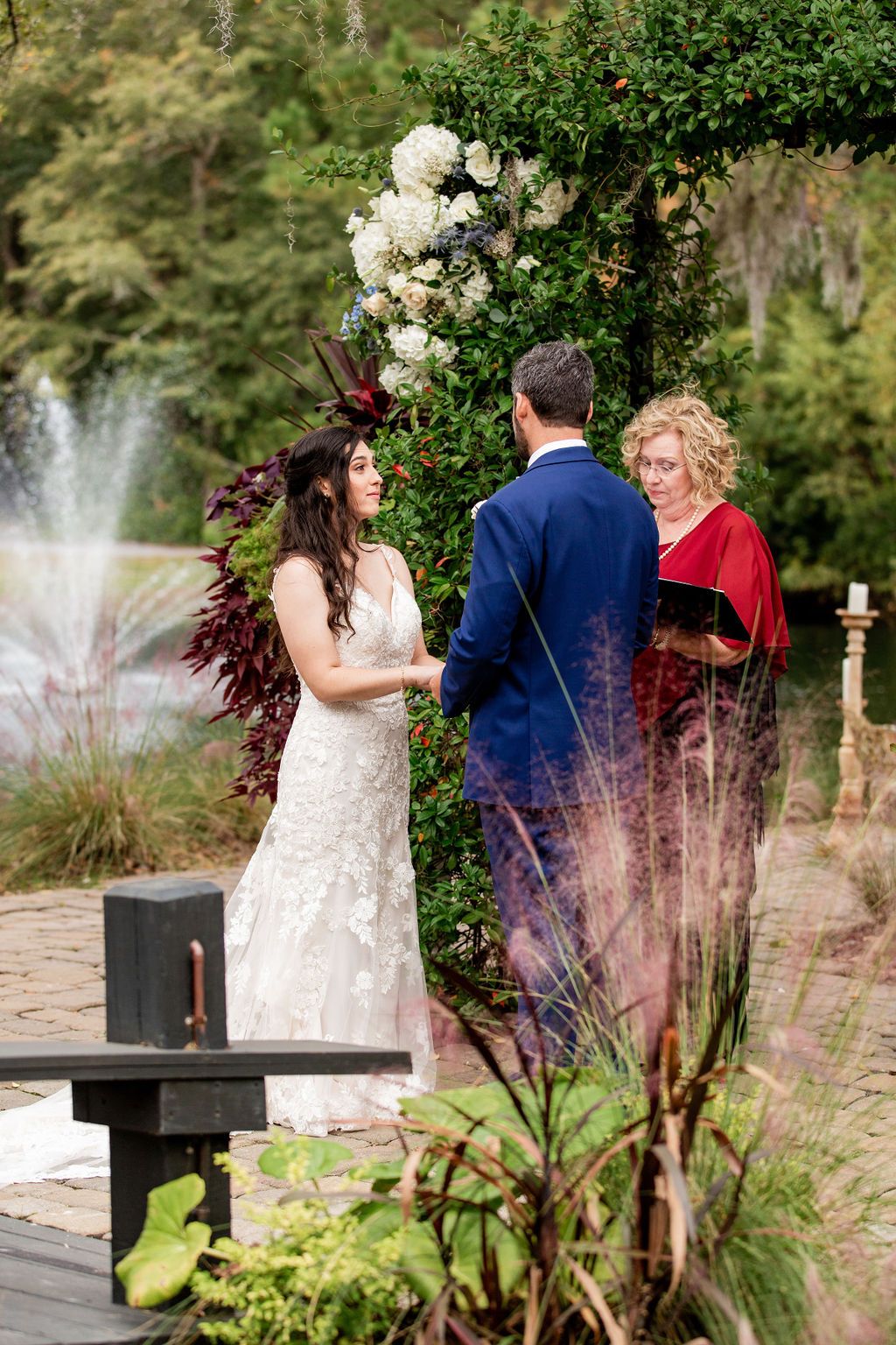 Wedding ceremony in a garden, with a bride and groom facing an officiant near flowers and a fountain.