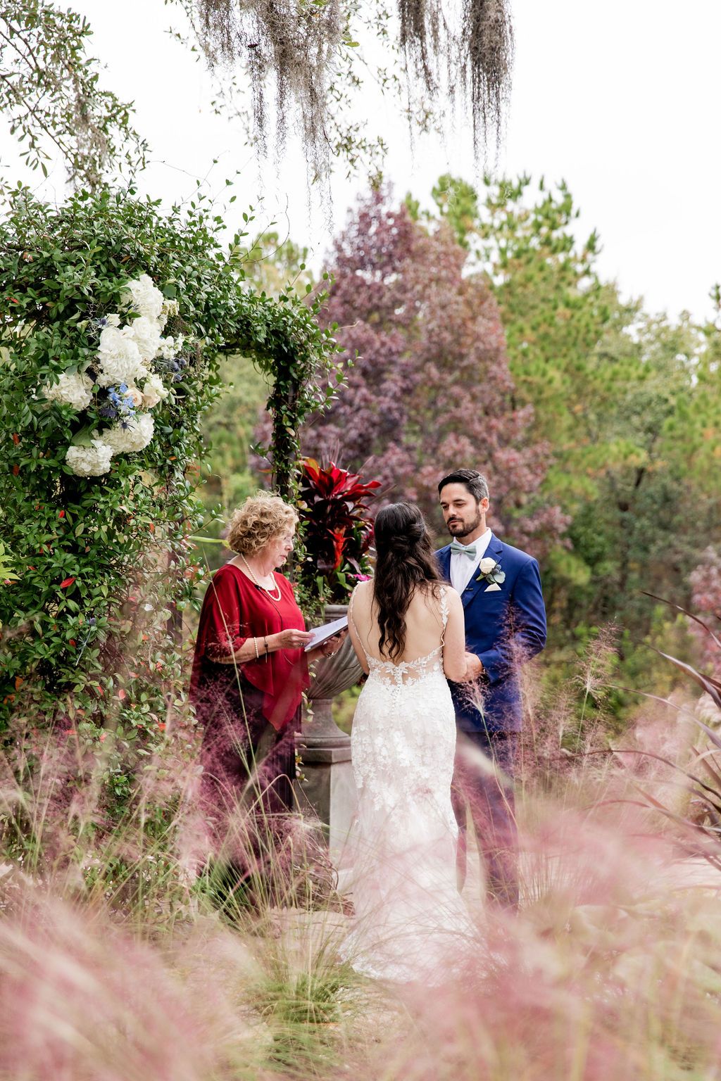 Three people in formal attire stand in a pink-flower garden, facing each other amid greenery.