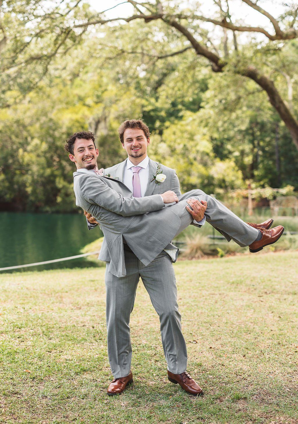 Two men in gray suits pose outdoors by a pond, one holding the other in his arms.