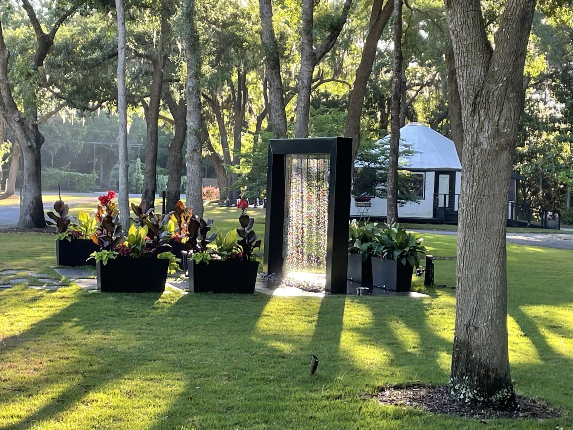Memorial garden with flower planters around a black water fountain under trees in a sunny park