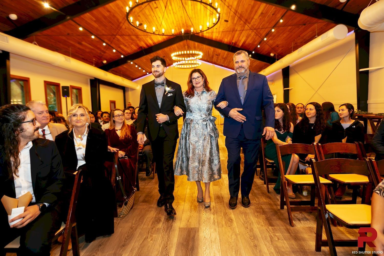 Wedding guests walk down the aisle in a decorated indoor venue, with the bride and groom in front.