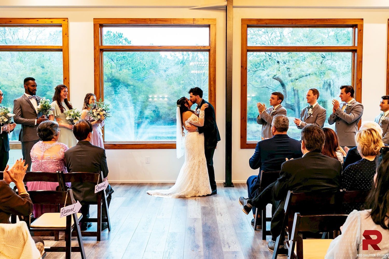 Wedding ceremony in a bright room with large windows, bride and groom at the altar, guests seated and clapping