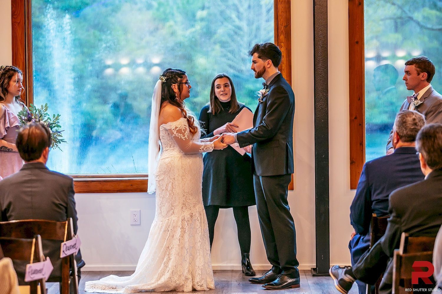 Wedding couple exchanging vows beside large windows, with officiant between them and guests seated nearby.