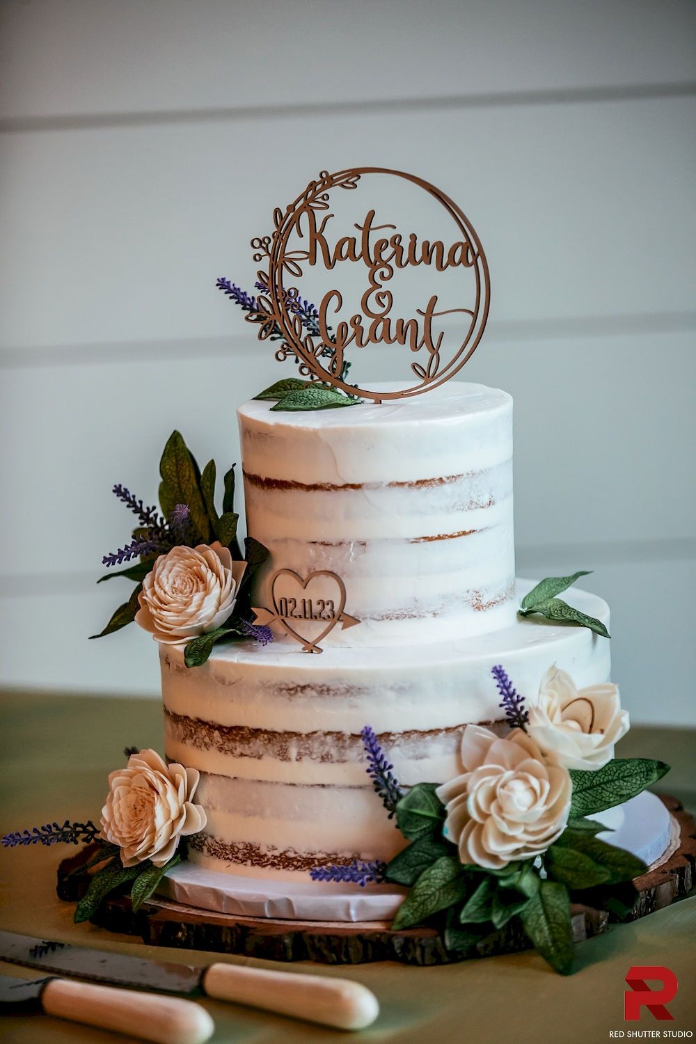 Rustic white tiered cake with cream flowers and gold topper on a wooden board.