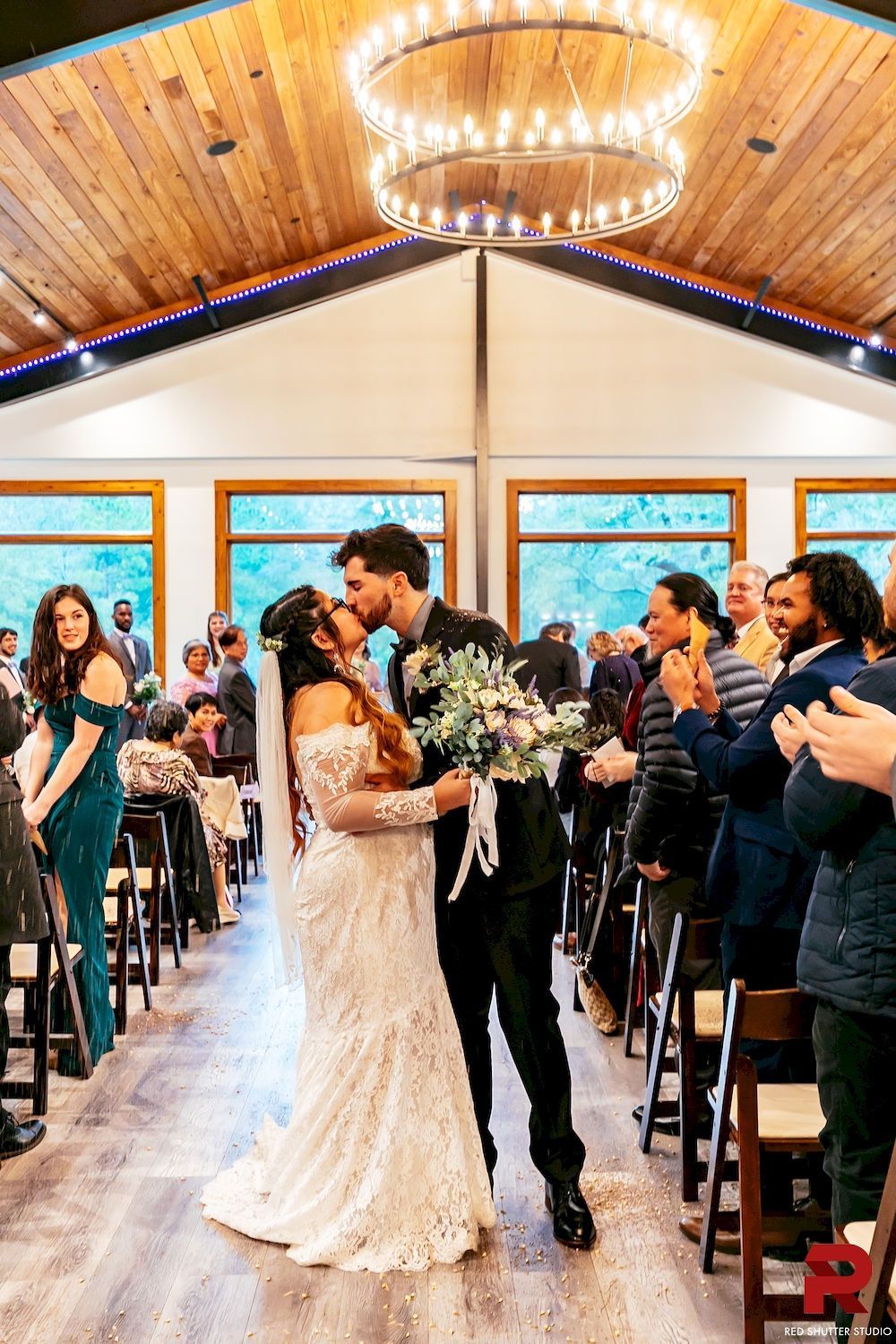 Bride and groom kiss in a warmly lit wedding aisle, guests standing and clapping nearby.
