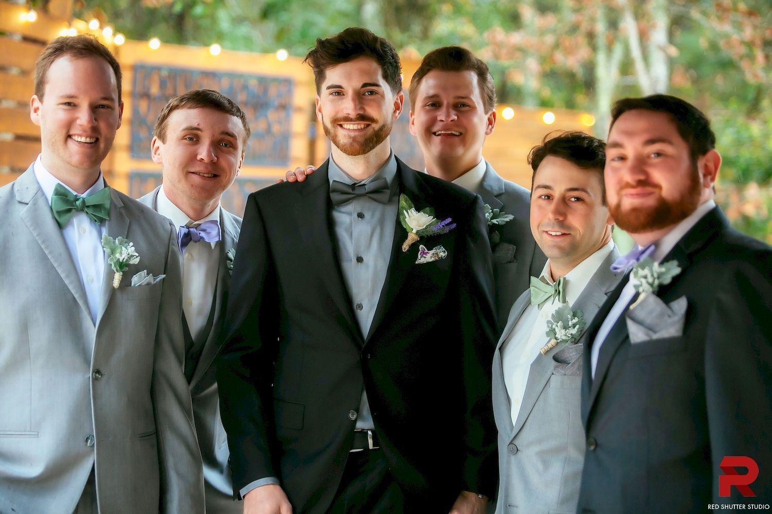 Five men in suits posing together outdoors, smiling in a decorated wedding setting