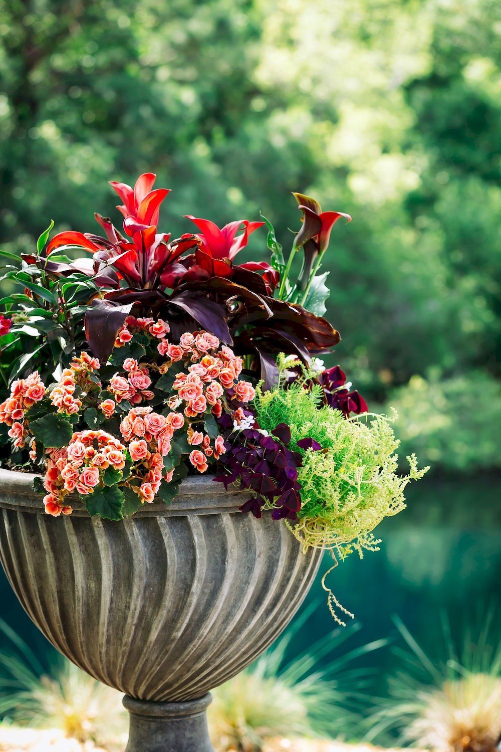 Decorative urn planter overflowing with red, pink, and purple flowers against a blurred green garden background