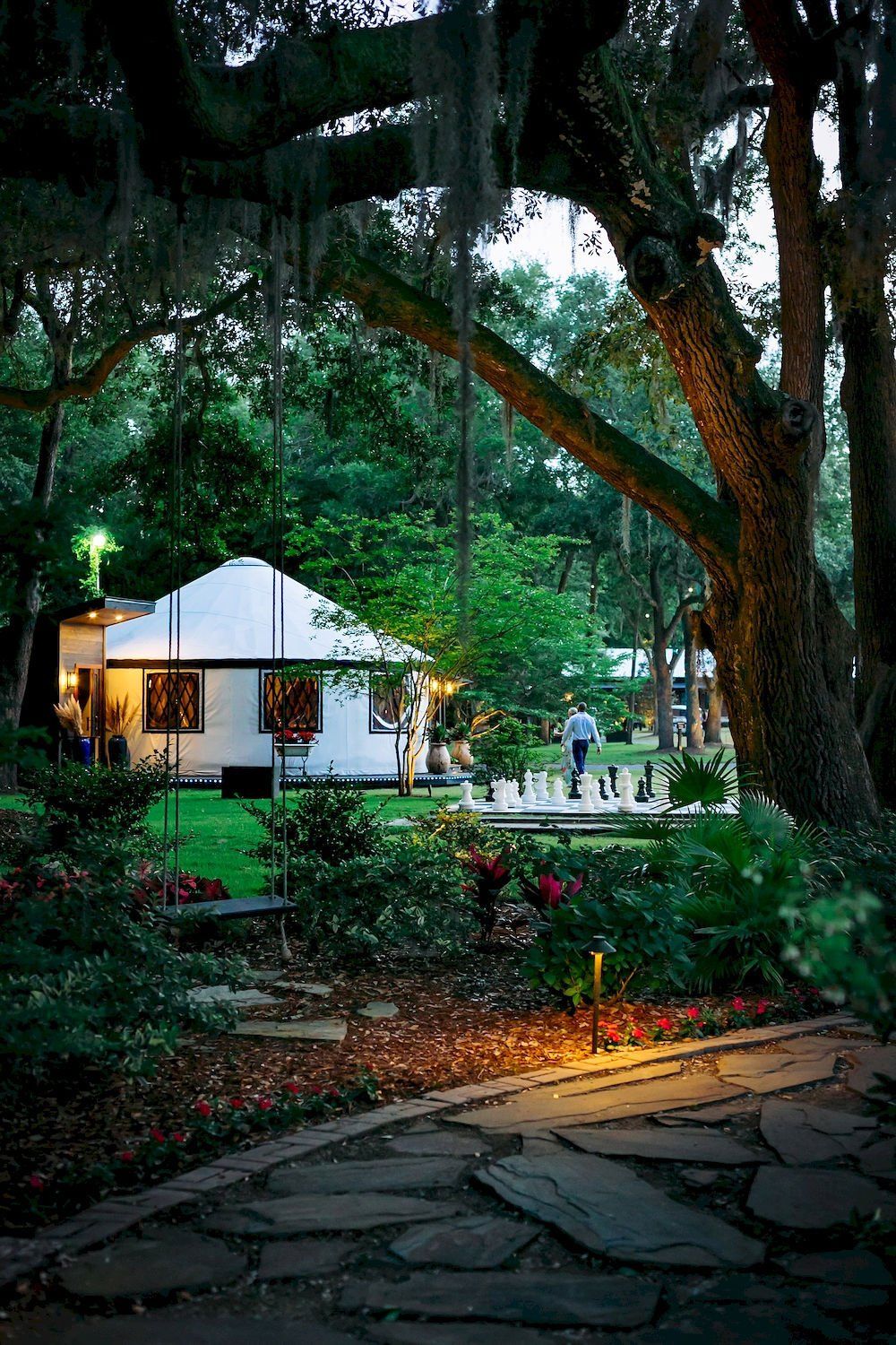 Lit garden resort path with a white tented building and large trees at dusk