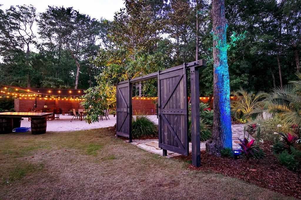 Open wooden gates in a lit garden at dusk, with string lights, trees, and a picnic table.