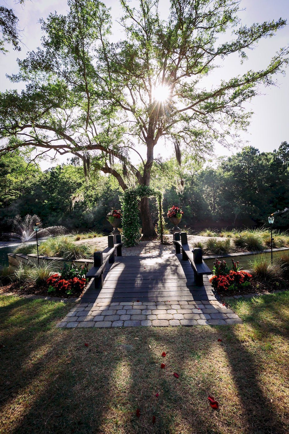 Sunlit park walkway lined with flowers and trees, with people standing in the distance