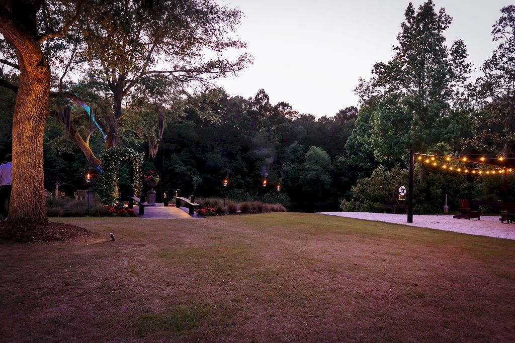 Dusk in a park with trees, a grassy field, and strings of warm lights along a path
