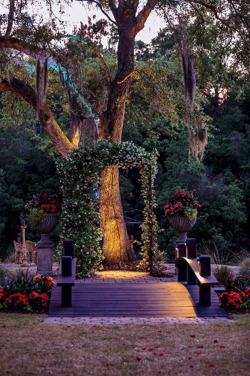 Lit wooden walkway leading to a vine-covered tree with hanging flower baskets in a garden at dusk