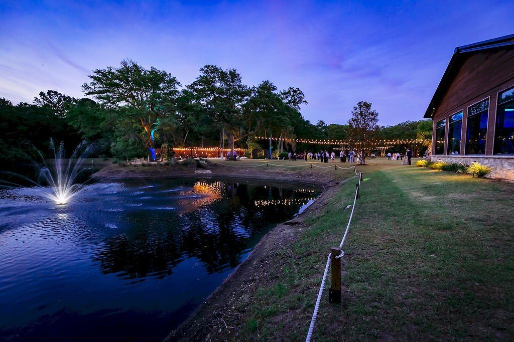 Dusk view of a lit pond with a fountain, trees, and a modern building beside a grassy shore