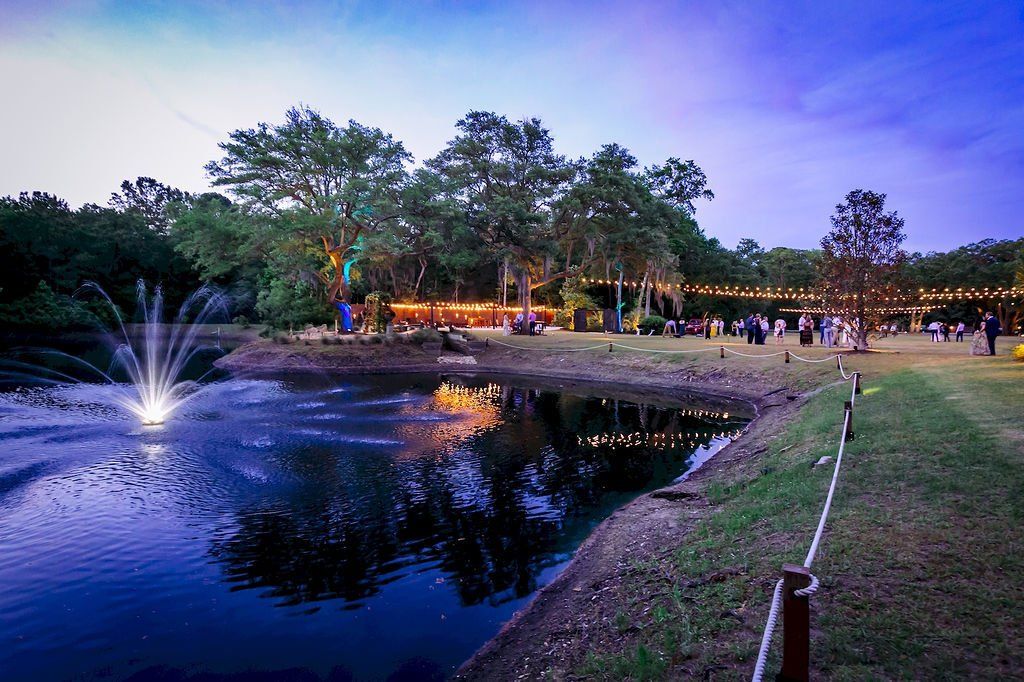 Park pond at dusk with a lit fountain, trees, and people along the shoreline