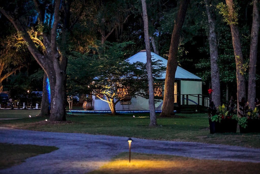 Park at dusk with lit pathway, trees, and a small house or pavilion in the background.