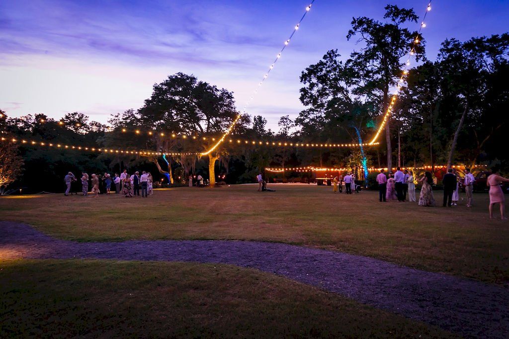 Outdoor evening gathering on a grassy field with string lights and groups of people under trees at sunset